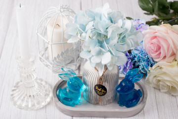 a bouquet of blue carnations with roses, Easter blue rabbits with a vase and a candlestick. On a wooden rustic background