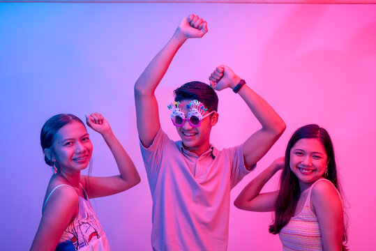 An extroverted young man dancing with two close female friends celebrating his birthday. Wearing funny novelty shades. Party scene lit with pink and blue neon colors.
