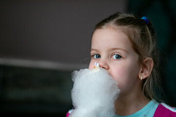 Portrait of amazing little girl with fair hair holding eating delicious white cotton candy...