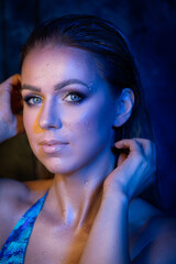 A portrait of a woman wearing a blue bathing suit, with droplets of water on her face, set against a blue background, and wearing waterproof makeup.
