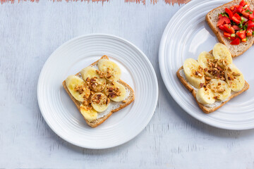 Sandwich breakfast has banana cream cheese almonds and tomato on white wood table.