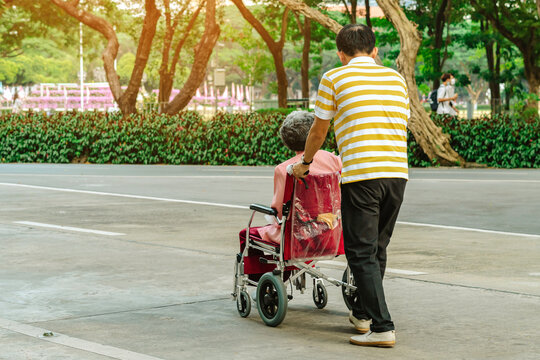 Back View Of Asian Elderly Man Walking With Disabled Elderly Woman Sitting In Wheelchair Outdoors Wearing Medical Masks. Man Pushes Old Lady In Wheelchair Through Park. Healthy Strong Medical Concept.