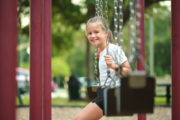 Young pretty happy smiling teenage small girl having fun on playground swings on summer vacations...