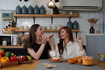 Beautiful woman and friend talking and using laptop at kitchen room. Girl friends couple living together at home.