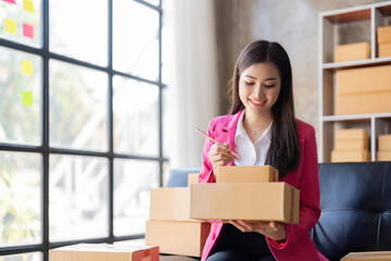 Young woman, an Asian online business owner, smiles, prepares parcel boxes and checks online orders...