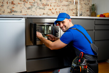 Young repairman in a blue uniform repairs and repairs a microwave oven with a screwdriver in a...