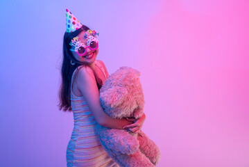 A young female birthday celebrant wearing a party hat and novelty glasses and holding a teddy bear....