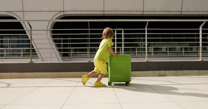 Boy is pushing green suitcase with all his might. Boy helps adult roll suitcase to waiting area at airport. Sympathetic boy is happy to help his parents with luggage. Young courageous assistant