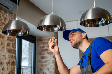 Male electrician changes the light bulbs in the lamp. A young man in overalls performs technical...