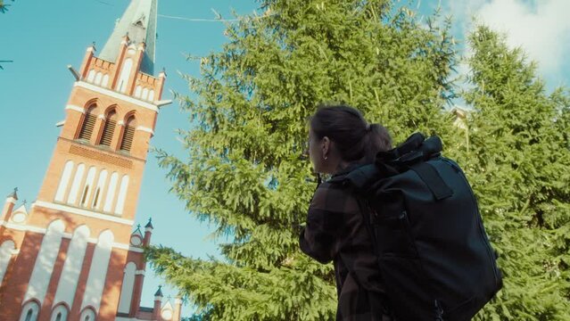 Close-up of female tourist with touring backpack is taking pictures of chatolic church with her camera. Woman photographer takes pictures of facade of old building