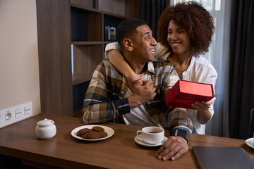 Cute multiracial wife giving gift box to husband at hotel breakfast