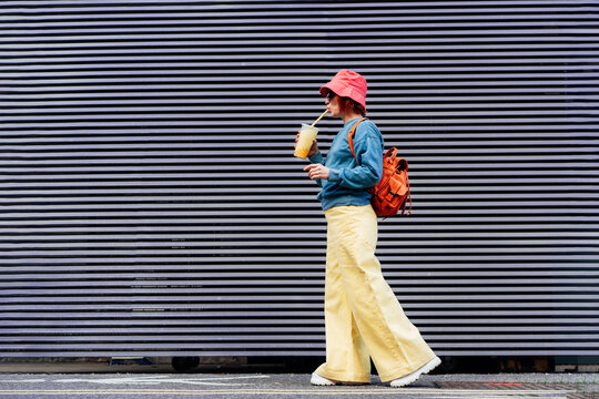 Stylish girl in bright clothes drinking sugar flavored tapioca bubble tea while walking near gray striped urban wall. Full length portrait of fashionable hipster girl. Street fashion. Copy space.
