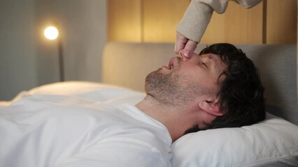 A heterosexual couple is in bed, a man is sleeping and hammering with his mouth open, while a tired woman, irritated by snoring, holds a man's nose