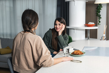Younger sister visiting her older sister in her new apartment she moved into after divorcing her abusive ex-husband. Two women sitting and talking at the kitchen counter drinking coffee and enjoy