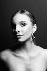 Fototapeta premium Beauty and make-up concept. Beautiful woman portrait with fancy make-up, classic hairstyle, naked shoulders and round earring standing in dark background and looking at camera. Black and white image
