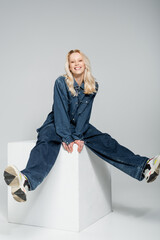 full length of happy woman in denim outfit and trendy sneakers sitting on white cube on grey.