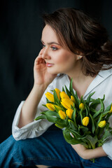 Spring portrait of young beautiful woman with blue eyes and curly hair with bouquet of yellow tulips on black background. Happy model in jeans and white shirt. Mothers Day.