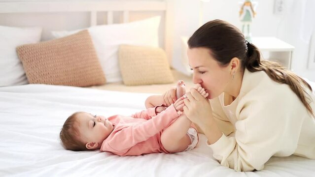 mom gently kisses her little daughter's legs at home on the bed and have fun together, family motherhood and love.