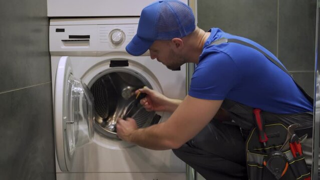 Installation or repair of a washing machine. A plumber in a blue uniform and a cap repairs a washing machine in the bathroom. 