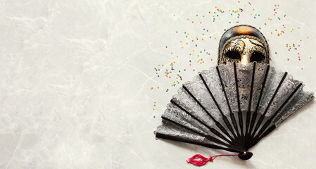 An elegant lace fan with Burano and vintage carnival mask on light marble table surrounded by colored confetti. Flat lay. Copy space for text. Toning. Close up.