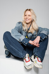 full length of happy blonde woman in blue denim outfit and trendy sneakers sitting on white cube...