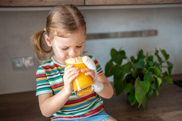 Little girl drinking fresh juice sitting on table in home kitchen