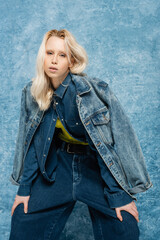 blonde woman in denim jacket posing while looking at camera near blue textured background.