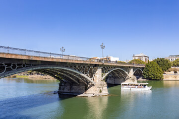 Fototapeta na wymiar Tourist boat crossing under El Puente de Isabel II, known as Puente de Triana, is a bridge located in Seville. Links the city center with the Triana neighborhood, crossing the Guadalquivir river