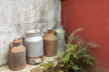 Rusted Old Metal Milk Jugs Containers Vats Sitting on a Stone Bench at a Farm Near Painted Red Wall