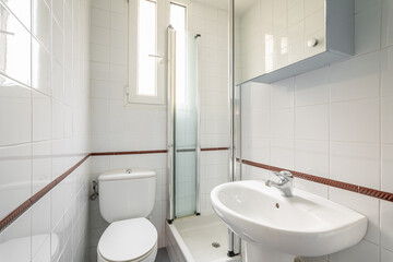 Luxury bathroom is lined with white ceramic tiles with shower toilet and sink with a narrow vertical windows for natural light. Concept of compact but comfortable bathrooms