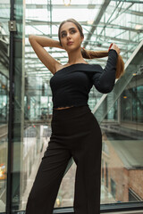 Beautiful young woman with a slender body in a fashion elegant business clothes with a top and trousers stands and fixes her hair in a modern office glass building