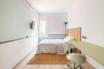 Narrow long room with bed, on opposite wall is TV for viewing watchable content. In corner is floor lamp with black shade for soft lighting at night. On floor is soft warm brown carpet.