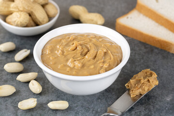 A bowl of peanut butter, toast, peanuts and a peanut butter knife on a gray background. American food.