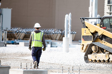 A construction worker walking on a job site with a skid steer in the background