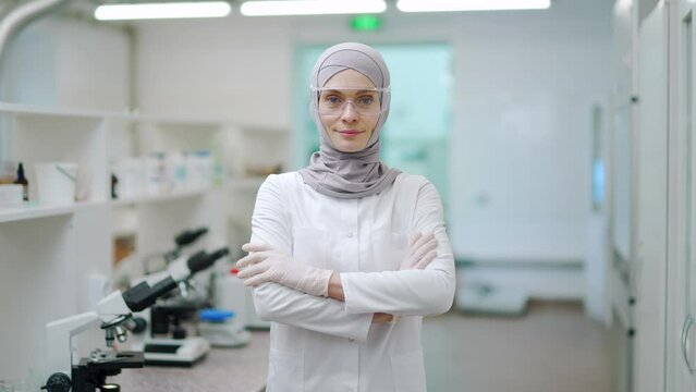Portrait of smiling muslim mature woman scientist researcher with protective glasses and gloves crossed arms and looking at the camera at microbiology pharmaceutical biochemistry laboratory hospital