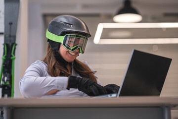 Crazy passionate skier sitting in an office with ski equipment and searching on her laptop next ski destination