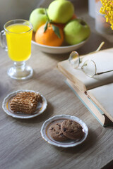 Cup of tea, plates with cookies, glass of orange juice, books, reading glasses, bowl of fruit and candles on the table. Selective focus.