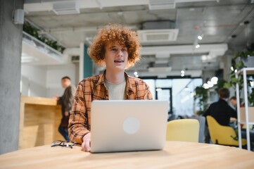 Portrait of Caucasian male freelancer in trendy apparel sitting at cafeteria table and doing remote work for programming design of public website, skilled software developer posing in coworking space.