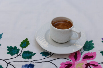 A cup of espresso coffee on a white tablecloth with patterns of flowers