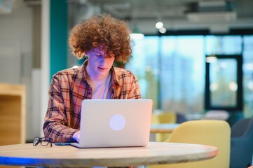 Portrait of Caucasian male freelancer in trendy apparel sitting at cafeteria table and doing remote work for programming design of public website, skilled software developer posing in coworking space.