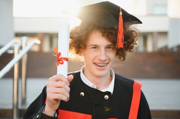 Happy smiling university graduate in mantle holding diploma in raised hand and expressing happiness...