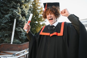 excited graduate student in gown with risen hands holding diploma.
