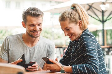 Obraz premium Young couple smiling while looking at their phone, sitting at a bar waiting for their drinks 
