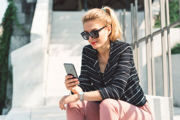 Fototapeta premium Woman in sunglasses looking at her phone while sitting on sun in the city