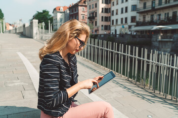 Fototapeta premium Beautiful woman with blonde hair looking down at the tablet while sitting on the sun outside int he city