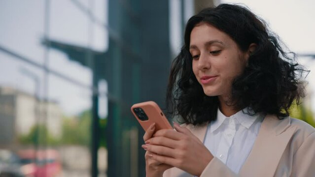 Beautiful Young Multi-Ethnic Curly Smiling Businesswoman in Formal Outfit Standing Near the Business Centre Using Smartphone. Close Up. People and Technology Business Concept