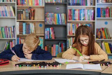 Two schoolchildren write and draw sitting at table in library. Classmates study in classroom.