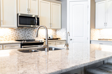 New modern faucet and kitchen sink with large island and granite countertops with bokeh background of microwave and oven stove with bright white cabints and nobody