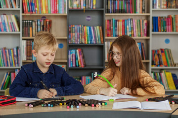 Fototapeta premium Two schoolchildren write and draw sitting at table in library. Classmates study in classroom.