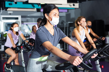 Men and women wearing protective masks ride stationary bike in a fitness club